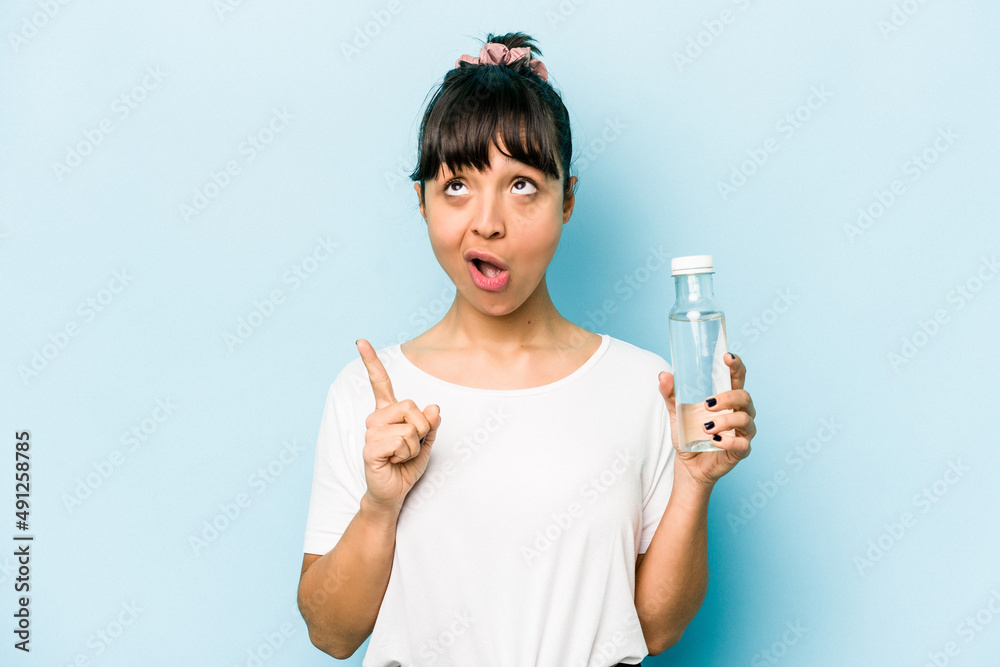 Young hispanic woman holding a bottle of water isolated on blue background pointing upside with opened mouth. Young hispanic woman holding a bottle of water isolated on blue background pointing upside with opened mouth.