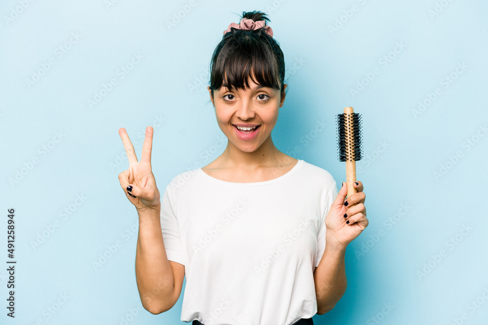 Young hispanic woman holding a brush hair isolated on blue background showing number two with fingers. Young hispanic woman holding a brush hair isolated on blue background showing number two with fingers.