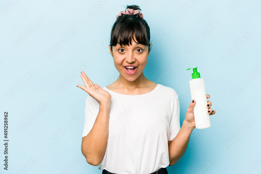 Young hispanic woman holding a body lotion isolated on blue background surprised and shocked.
