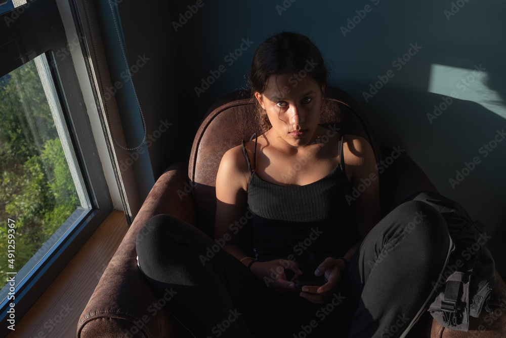 A young woman sits on a sofa near a window with phone in her hand sunny day