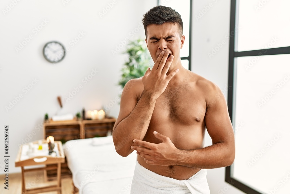 Obraz premium Young hispanic man standing shirtless at spa center bored yawning tired covering mouth with hand. restless and sleepiness.