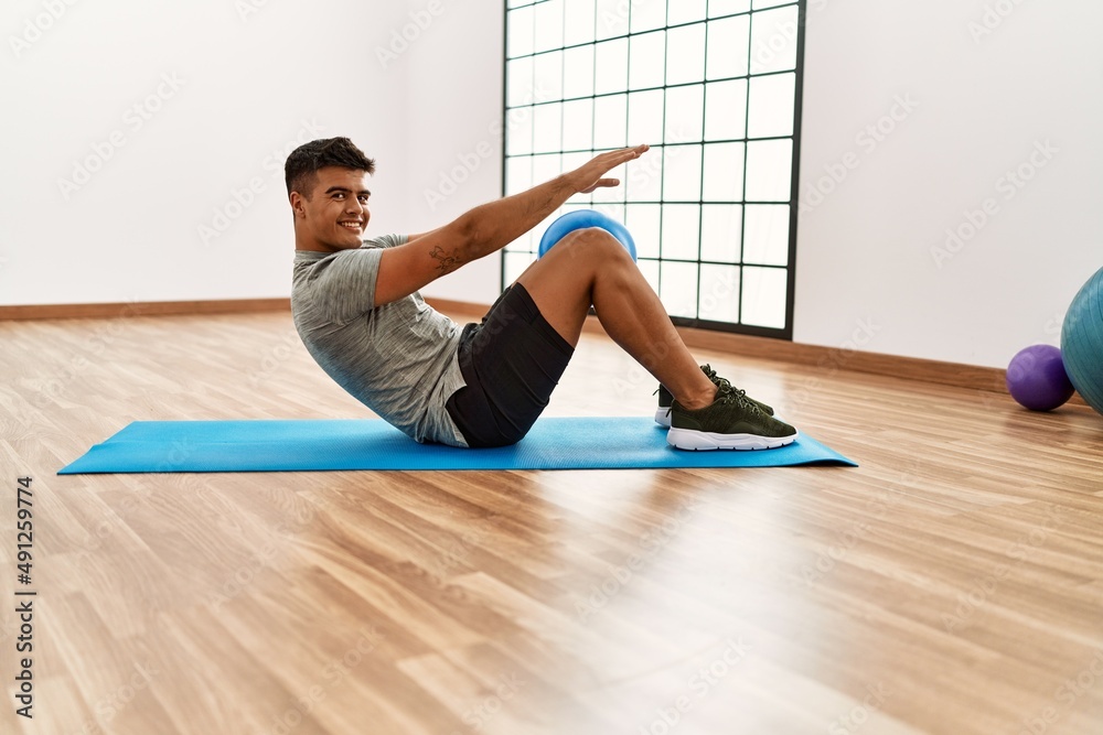 Young hispanic man smiling confident training abs exercise using ball at sport center