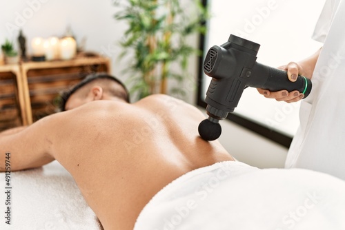 Young hispanic man relaxed having back massage using percussion pistol at beauty center