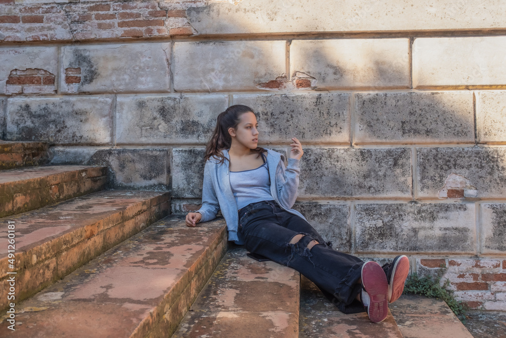 Young woman sits on grey stairs dressed in ripped jeans brick wall behind Young woman sits on grey stairs dressed in ripped jeans brick wall behind