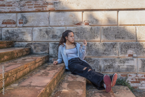 Young woman sits on grey stairs dressed in ripped jeans brick wall behind