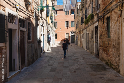 Young tourists walk on a narrow alley of Venice old brick houses on both sides