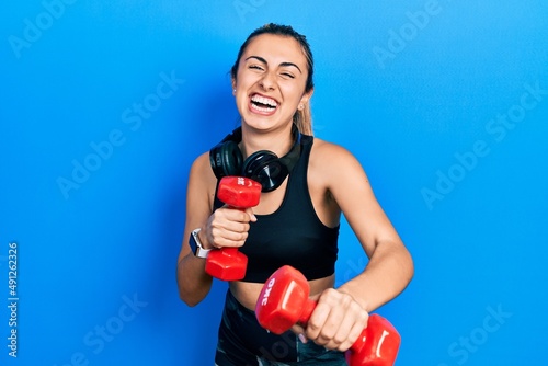 Beautiful hispanic woman wearing sportswear using dumbbells smiling and laughing hard out loud because funny crazy joke.