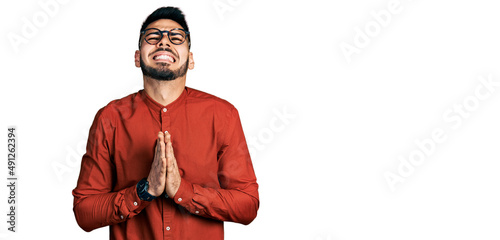 Young hispanic man with beard wearing business shirt and glasses begging and praying with hands together with hope expression on face very emotional and worried. begging. photo