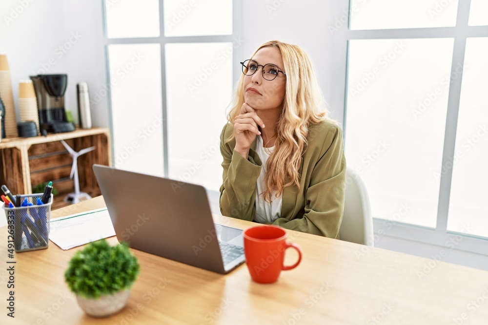 Beautiful blonde woman working at the office with laptop with hand on chin thinking about question, pensive expression. smiling with thoughtful face. doubt concept. Beautiful blonde woman working at the office with laptop with hand on chin thinking about question, pensive expression. smiling with thoughtful face. doubt concept.