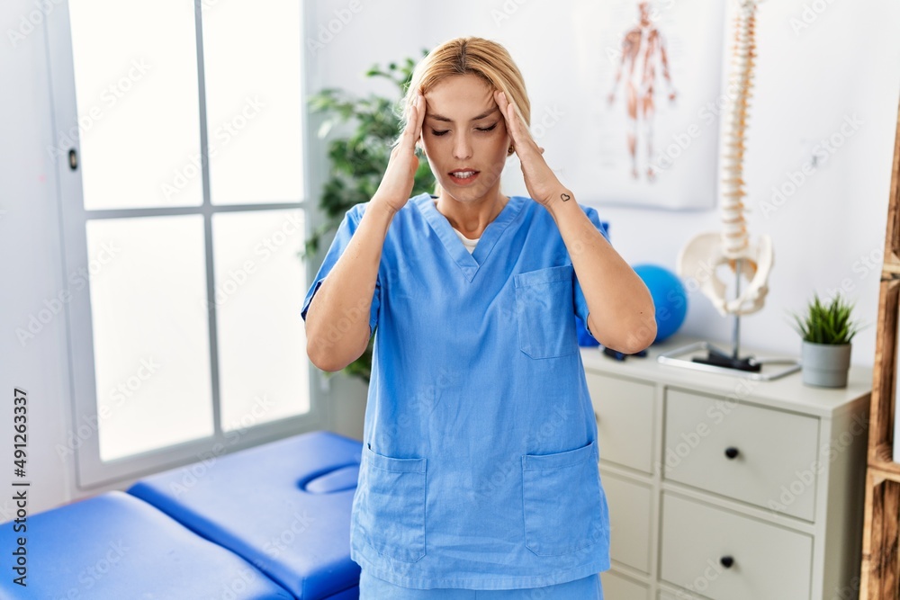 Beautiful blonde physiotherapist woman working at pain recovery clinic with hand on head for pain in head because stress. suffering migraine. Beautiful blonde physiotherapist woman working at pain recovery clinic with hand on head for pain in head because stress. suffering migraine.