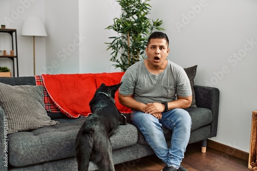Young latin man and dog sitting on the sofa at home afraid and shocked with surprise and amazed expression, fear and excited face. © Krakenimages.com