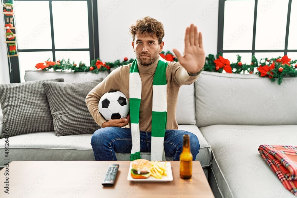 Young hispanic man football hooligan holding ball at home with open hand doing stop sign with serious and confident expression, defense gesture