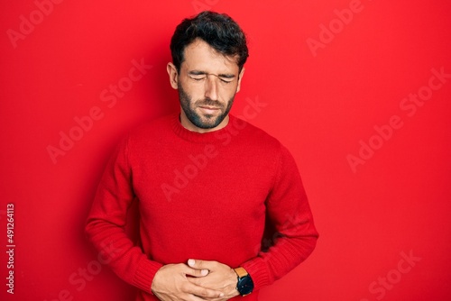 Handsome man with beard wearing casual red sweater with hand on stomach because indigestion, painful illness feeling unwell. ache concept. © Krakenimages.com