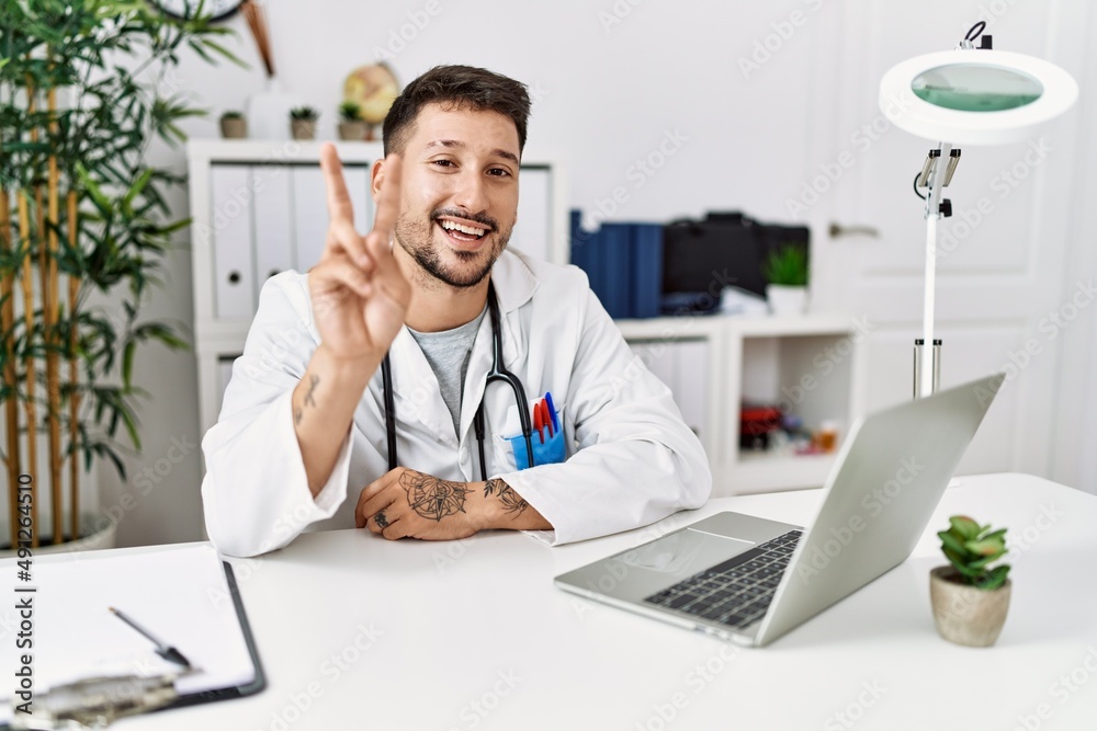 Young doctor working at the clinic using computer laptop smiling with happy face winking at the camera doing victory sign. number two. Young doctor working at the clinic using computer laptop smiling with happy face winking at the camera doing victory sign. number two.