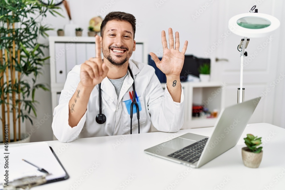 Young doctor working at the clinic using computer laptop showing and pointing up with fingers number six while smiling confident and happy.