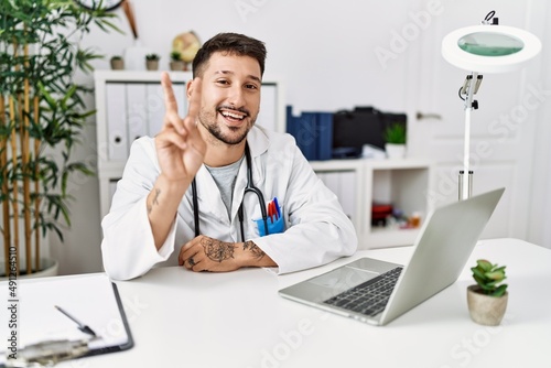 Young doctor working at the clinic using computer laptop smiling with happy face winking at the camera doing victory sign. number two.