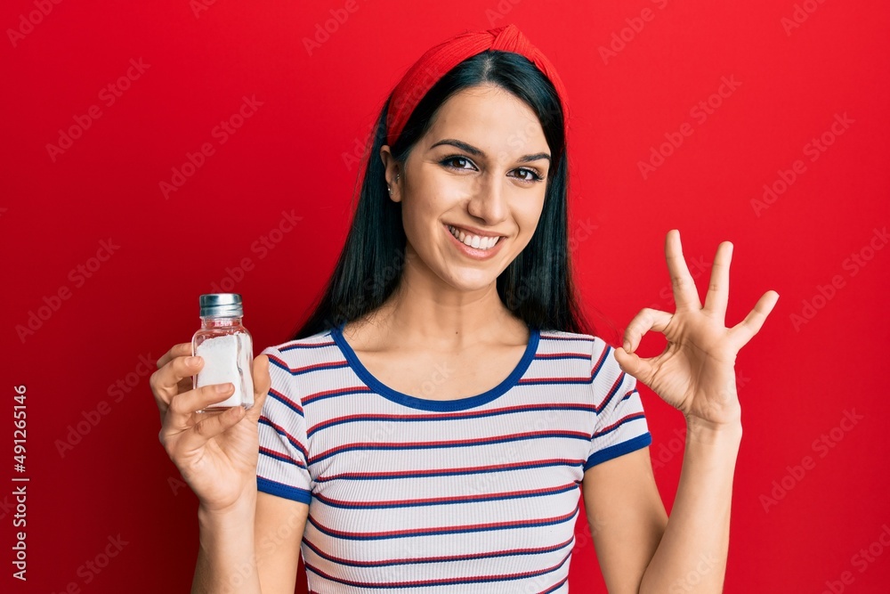 Young hispanic woman holding salt shaker doing ok sign with fingers, smiling friendly gesturing excellent symbol Young hispanic woman holding salt shaker doing ok sign with fingers, smiling friendly gesturing excellent symbol