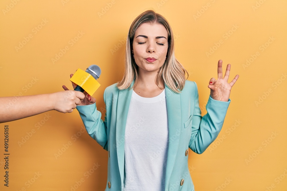 Beautiful caucasian woman being interviewed by reporter holding microphone relax and smiling with eyes closed doing meditation gesture with fingers. yoga concept. Beautiful caucasian woman being interviewed by reporter holding microphone relax and smiling with eyes closed doing meditation gesture with fingers. yoga concept.
