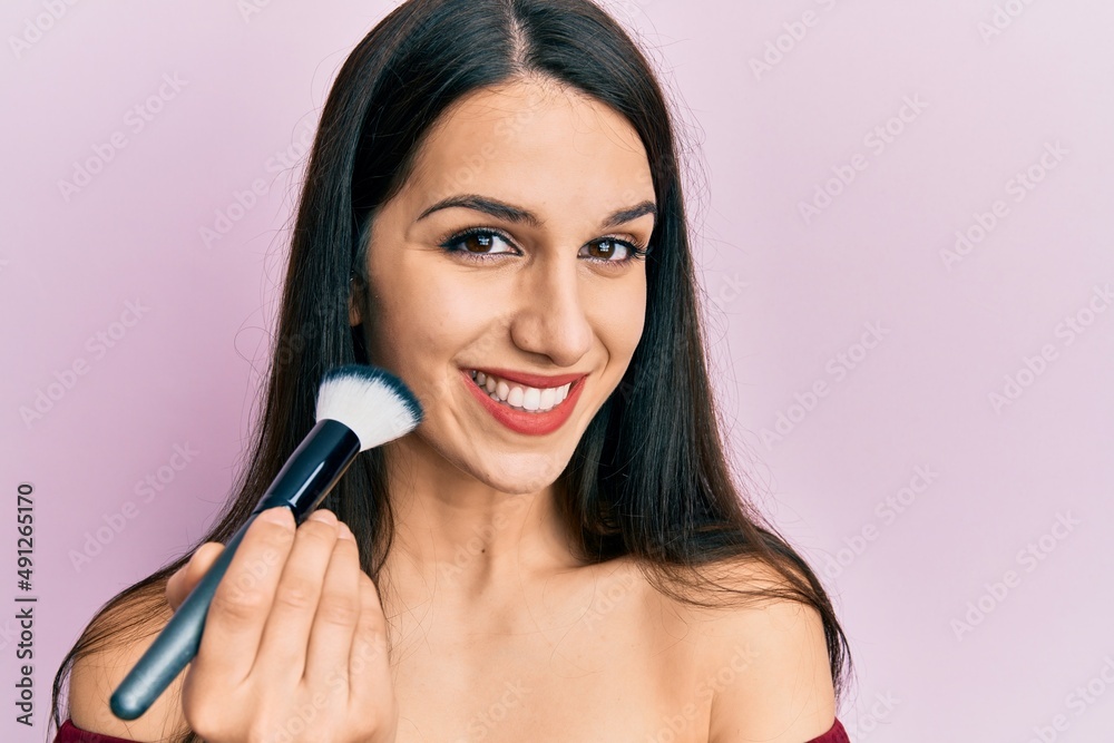 Young hispanic woman holding makeup brush and blush looking positive and happy standing and smiling with a confident smile showing teeth Young hispanic woman holding makeup brush and blush looking positive and happy standing and smiling with a confident smile showing teeth
