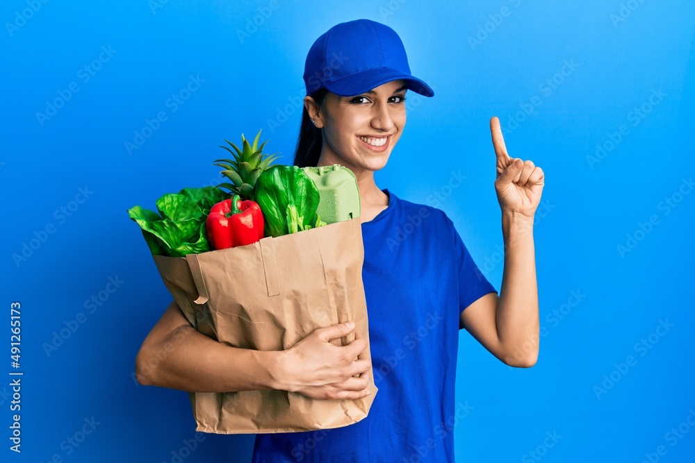 Young hispanic woman wearing courier uniform with groceries from supermarket smiling with an idea or question pointing finger with happy face, number one Young hispanic woman wearing courier uniform with groceries from supermarket smiling with an idea or question pointing finger with happy face, number one
