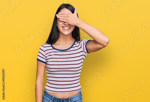 Young hispanic girl wearing casual striped t shirt smiling and laughing with hand on face covering eyes for surprise. blind concept.