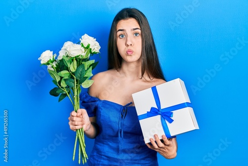 Young brunette teenager holding anniversary present and bouquet of flowers puffing cheeks with funny face. mouth inflated with air, catching air.
