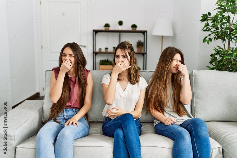 Group of three hispanic girls sitting on the sofa at home smelling something stinky and disgusting, intolerable smell, holding breath with fingers on nose. bad smell Group of three hispanic girls sitting on the sofa at home smelling something stinky and disgusting, intolerable smell, holding breath with fingers on nose. bad smell