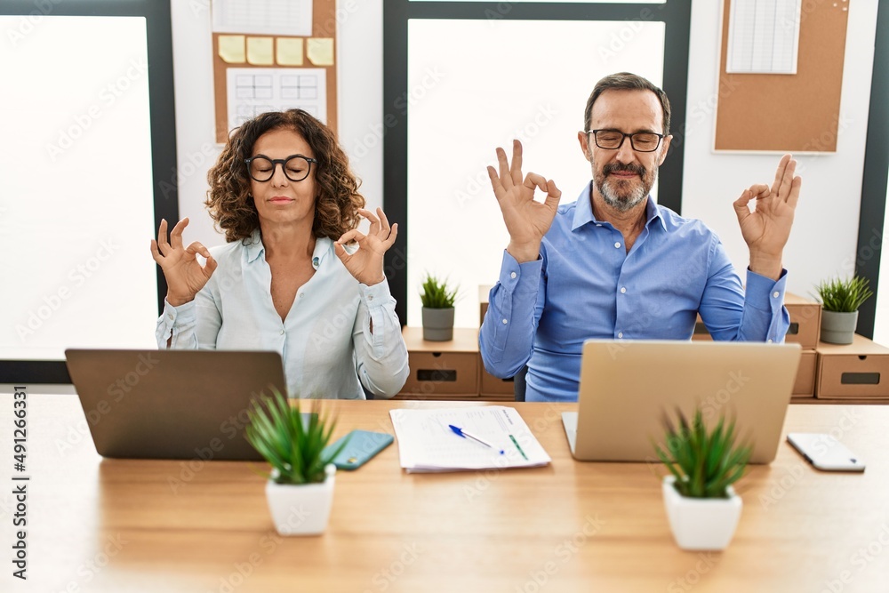 Fototapeta premium Middle age hispanic woman and man sitting with laptop at the office relax and smiling with eyes closed doing meditation gesture with fingers. yoga concept.