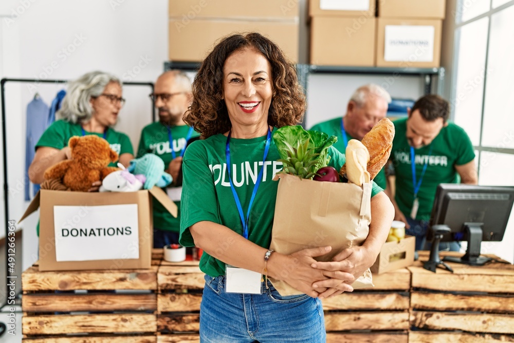 Group of middle age volunteers working at charity center. Woman smiling happy and holding paper bag with food to donate.