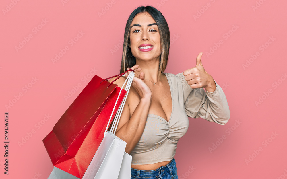 Young hispanic woman holding shopping bags smiling happy and positive, thumb up doing excellent and approval sign Young hispanic woman holding shopping bags smiling happy and positive, thumb up doing excellent and approval sign