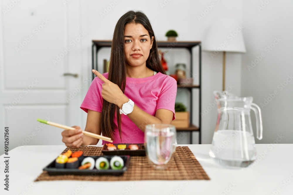 Young brunette woman eating sushi using chopsticks pointing with hand finger to the side showing advertisement, serious and calm face Young brunette woman eating sushi using chopsticks pointing with hand finger to the side showing advertisement, serious and calm face
