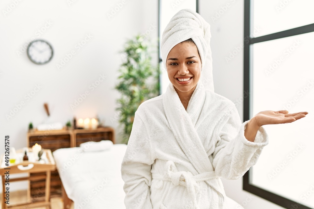 Young brunette woman wearing towel and bathrobe standing at beauty center smiling cheerful presenting and pointing with palm of hand looking at the camera.