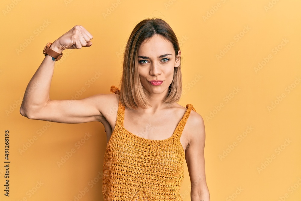 Young caucasian blonde woman wearing casual yellow t shirt strong person showing arm muscle, confident and proud of power Young caucasian blonde woman wearing casual yellow t shirt strong person showing arm muscle, confident and proud of power
