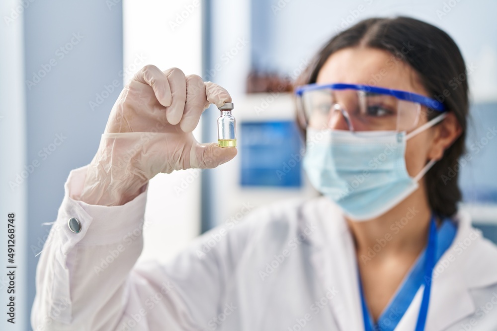 Young hispanic woman wearing scientist uniform and medical mask holding vaccine dose at laboratory