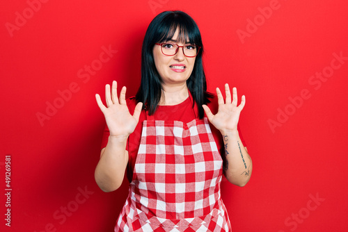 Young hispanic woman wearing cook apron and glasses afraid and terrified with fear expression stop gesture with hands, shouting in shock. panic concept. © Krakenimages.com