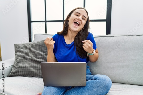 Young brunette woman using laptop at home very happy and excited doing winner gesture with arms raised, smiling and screaming for success. celebration concept.