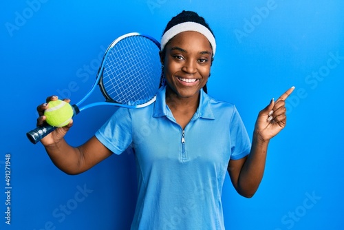 African american woman with braided hair playing tennis holding racket and ball smiling happy pointing with hand and finger to the side © Krakenimages.com