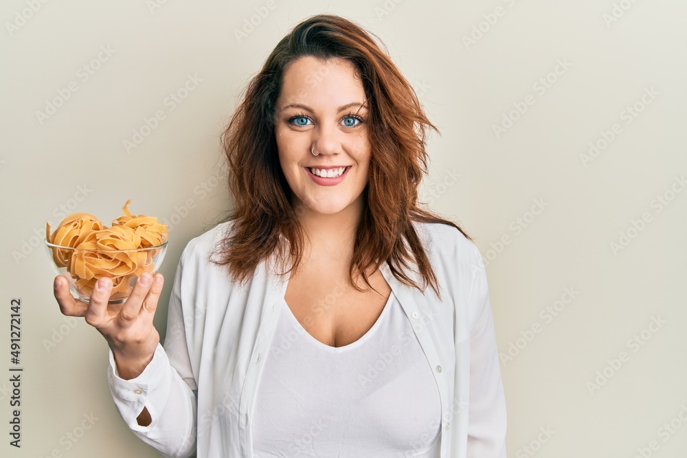 Young caucasian woman holding bowl of uncooked pasta looking positive and happy standing and smiling with a confident smile showing teeth