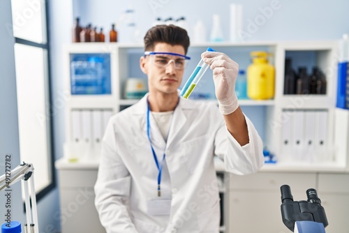 Young hispanic man wearing scientist uniform holding test tubes at laboratory