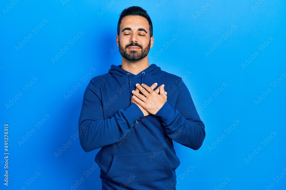 Handsome hispanic man with beard wearing casual sweatshirt smiling with hands on chest, eyes closed with grateful gesture on face. health concept. Handsome hispanic man with beard wearing casual sweatshirt smiling with hands on chest, eyes closed with grateful gesture on face. health concept.