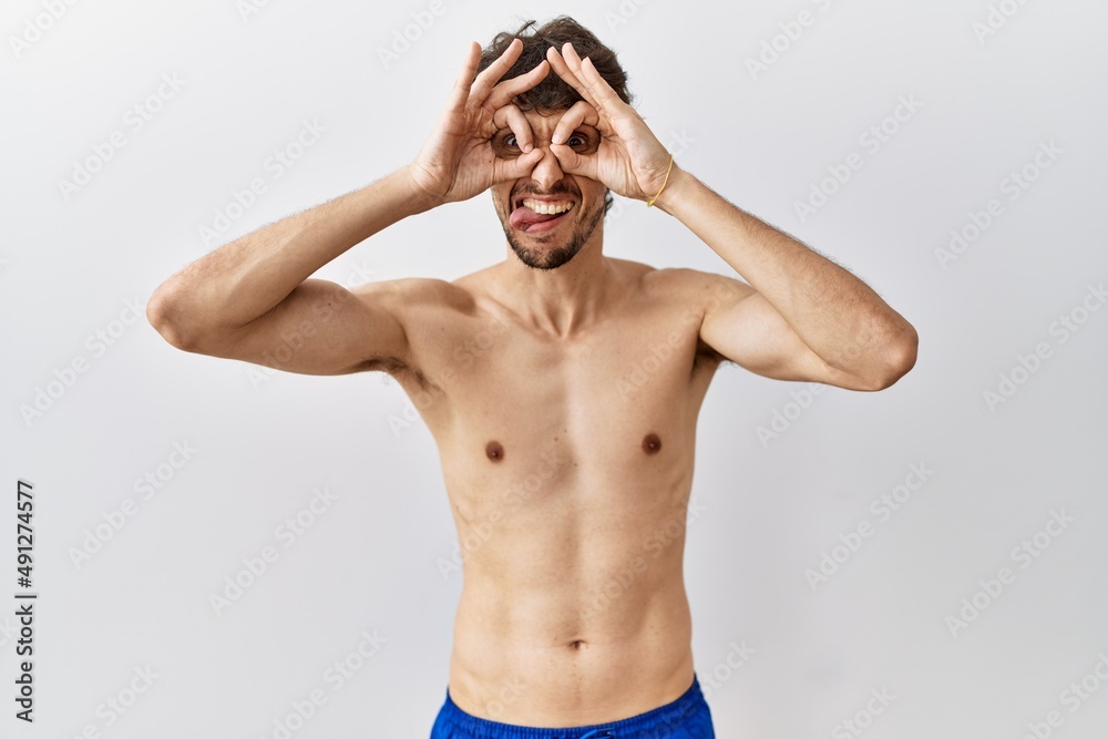 Fototapeta premium Young hispanic man standing shirtless over isolated, background doing ok gesture like binoculars sticking tongue out, eyes looking through fingers. crazy expression.