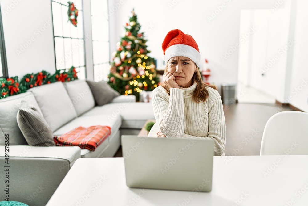 Middle age woman wearing santa claus hat using laptop looking stressed and nervous with hands on mouth biting nails. anxiety problem. Middle age woman wearing santa claus hat using laptop looking stressed and nervous with hands on mouth biting nails. anxiety problem.