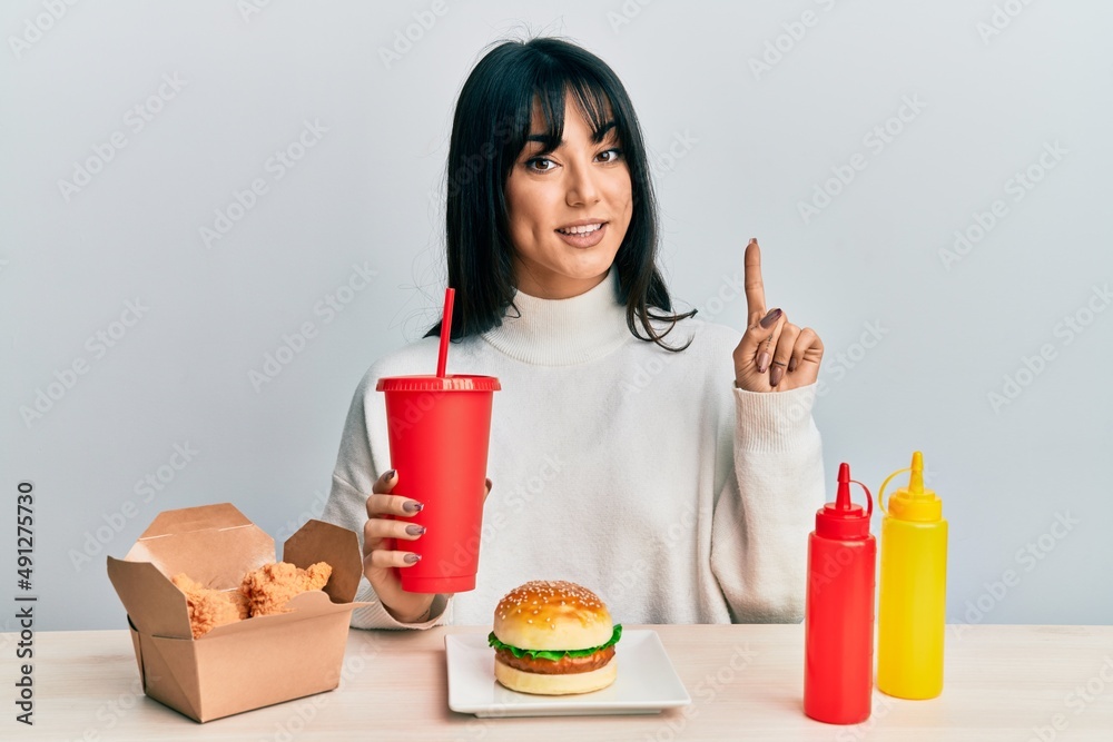 Young brunette woman with bangs eating a tasty classic burger with ketchup and mustard smiling with an idea or question pointing finger with happy face, number one Young brunette woman with bangs eating a tasty classic burger with ketchup and mustard smiling with an idea or question pointing finger with happy face, number one