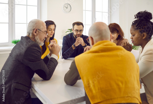 Diverse people praying together. Group of different religious multiracial multiethnic males and females sitting around table and saying prayer of gratitude, thanking God for His amazing grace