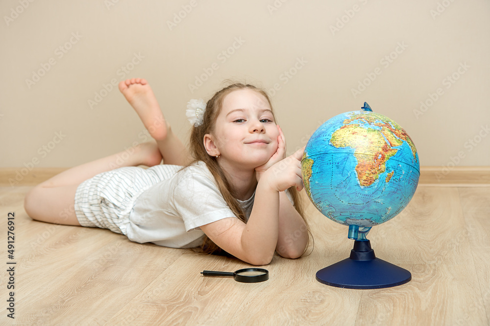cute little girl lies on the floor next to a globe and a magnifying glass and points with her finger where she would like to go travel, travel concept, travel choice, dreams and plans education