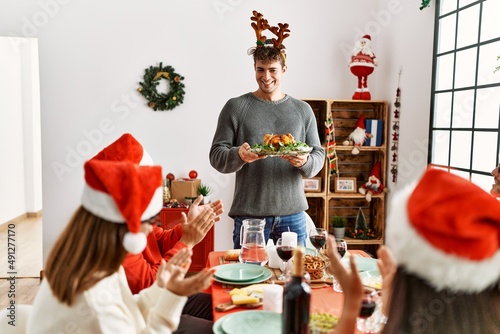 Group of people meeting clapping and sitting on the table. Man standing and holding roasted turkey celebrating Christmas at home.