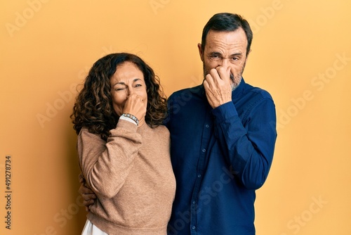 Middle age hispanic couple wearing casual clothes smelling something stinky and disgusting, intolerable smell, holding breath with fingers on nose. bad smell