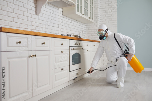 Pest control home service guy fighting parasites in the house. Exterminator in mask, goggles and PPE suit spraying poisonous gas or liquid from sprayer bottle on floor and cupboard in kitchen interior photo