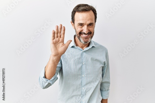 Middle age hispanic man with beard standing over isolated background waiving saying hello happy and smiling, friendly welcome gesture