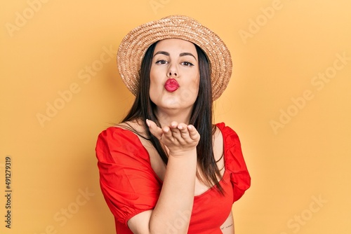 Young hispanic woman wearing summer hat looking at the camera blowing a kiss with hand on air being lovely and sexy. love expression.